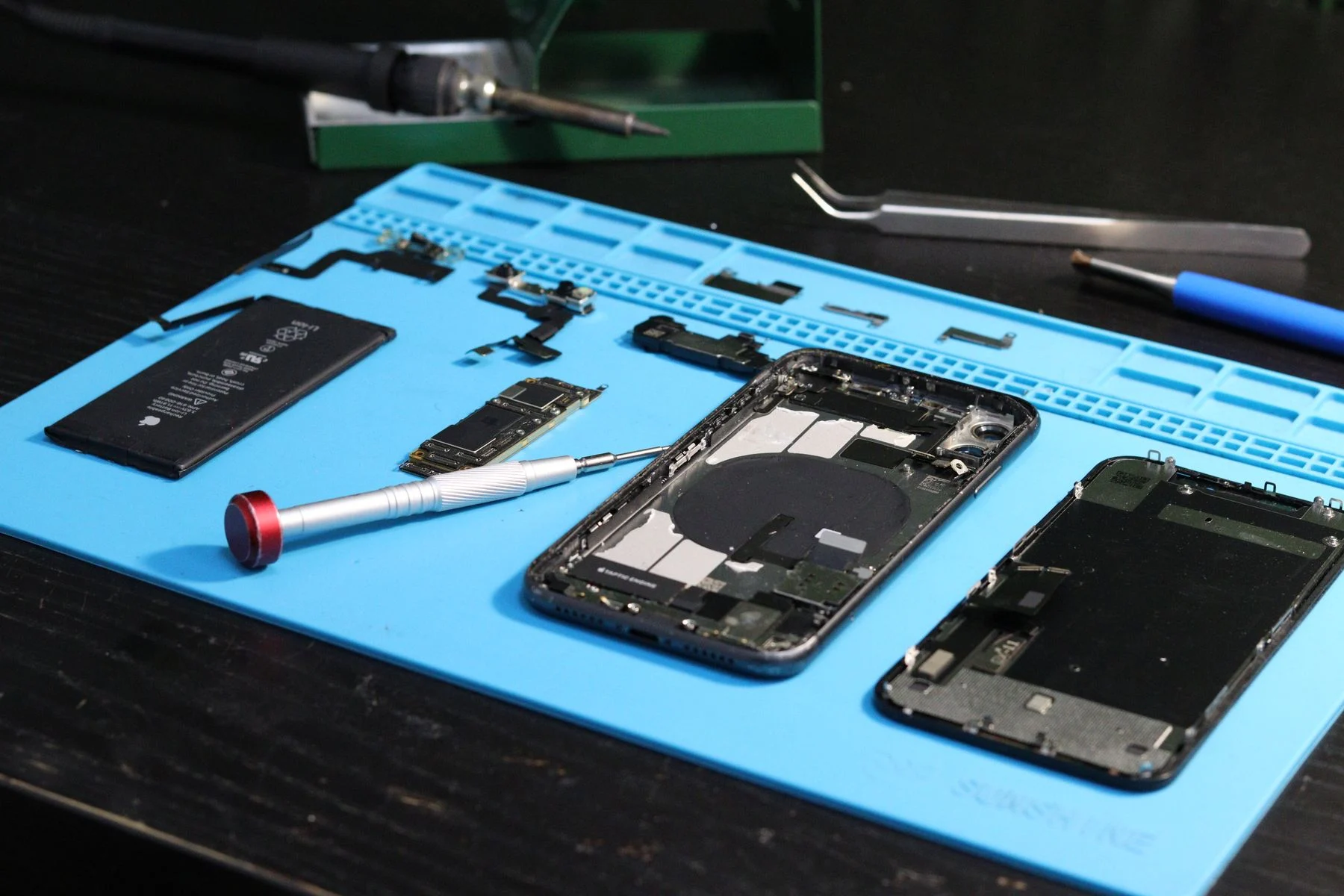 Zerlegtes iPhone mit Akku, Platine und Werkzeug auf einer blauen Reparaturmatte